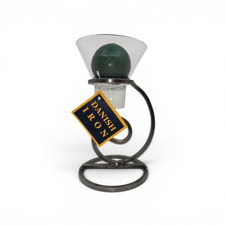 A dark green ball candle in a clear glass Leif cup rests on Talla Imports’ Danish Iron candle holder with a tall curl base; a yellow and blue “Danish Iron” tag hangs from the stand.