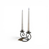 The Talla Imports Candle Holder: Danish Iron Double Curls holds two white taper candles in curled black iron arms, with a small rectangular tag attached. The holder is hand forged and set against a plain white background.