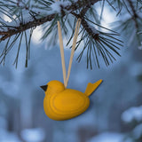The Swallings Wooden Yellow Swedish Bird Hanging Ornament, made in Sweden, hangs from a pine branch by two strings against a snowy, blurred outdoor background.