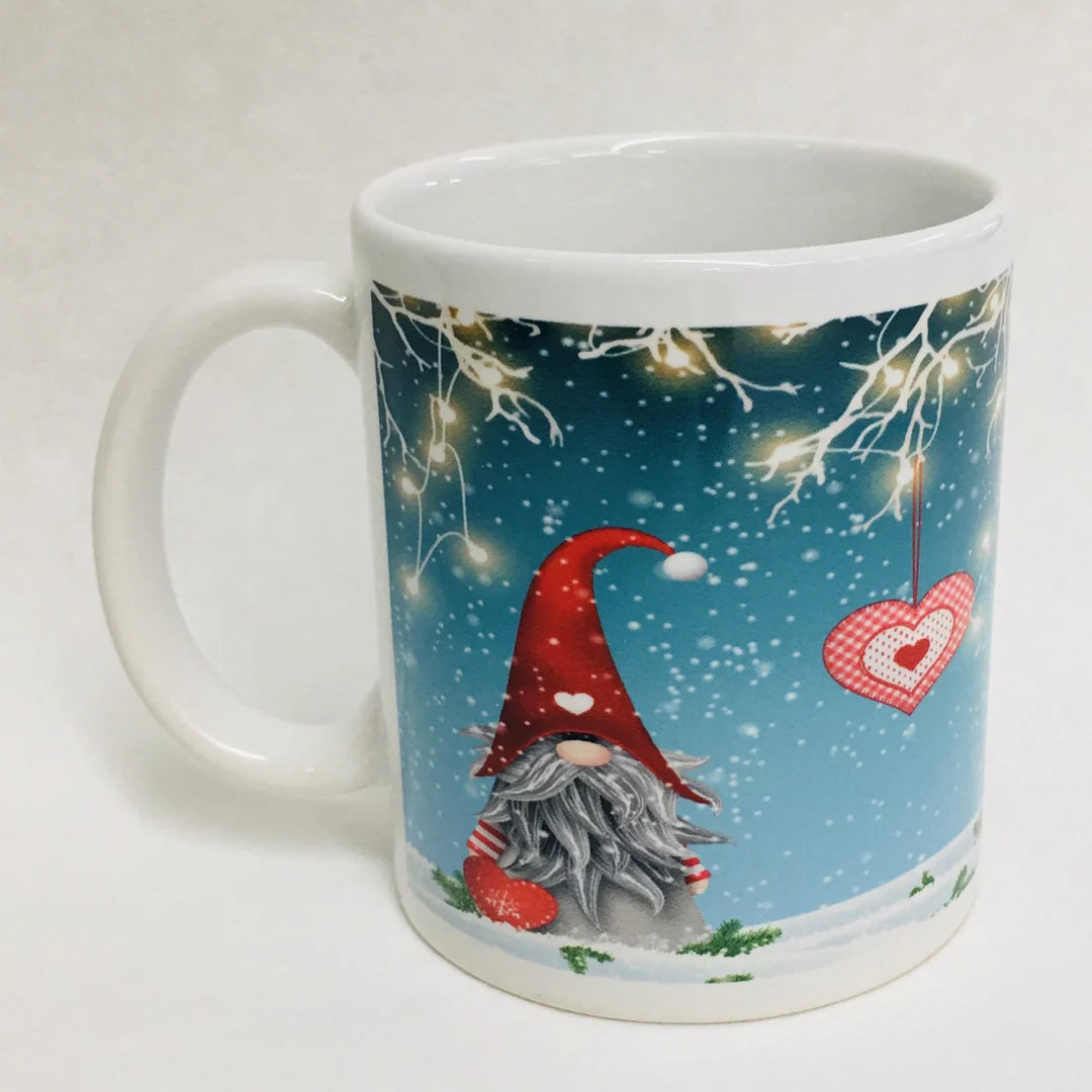 The Mug: Gnome with lights coffee mug is a white ceramic mug featuring a red-hatted gnome, snowy scene, lit tree branches, and heart ornament on a blue background—ideal for cozy holiday mornings.