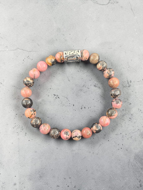 Bracelet featuring an Algiz rune charm, pink and brown marbled stones, and one ornate silver bead, shown on a gray stone background.