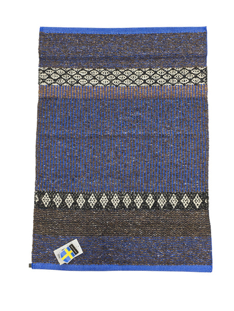 The Horredsmattan Mixed Forest Cobalt rug is a rectangular Swedish cotton/vinyl rug with blue, brown, and black patterns, white geometric designs, and a small Swedish flag tag in the bottom left corner.