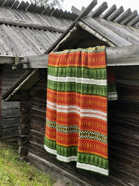 The Mora Blanket – 100% Norwegian Lamb’s Wool by Kerstin Landström Design, featuring colorful patterns, hangs over the wooden roof of a rustic log cabin.