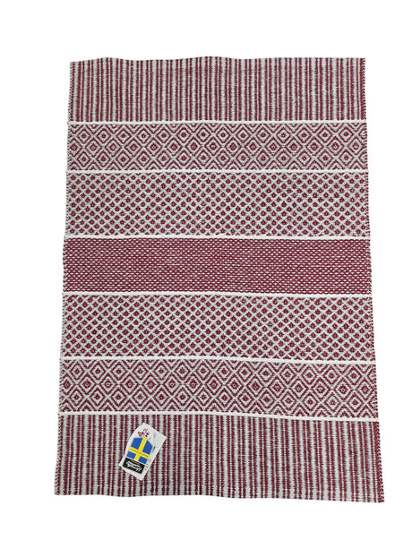 The Vinyl Rug: Horredsmattan - Alfie - Red is a rectangular Swedish vinyl rug featuring red and white geometric patterns and a corner tag with the Swedish flag.