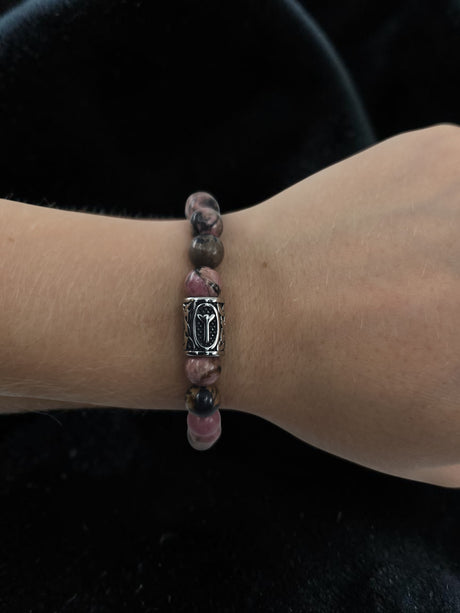A hand wearing a beaded bracelet with pink and brown stones and a silver Algiz rune charm, shown against a black background.