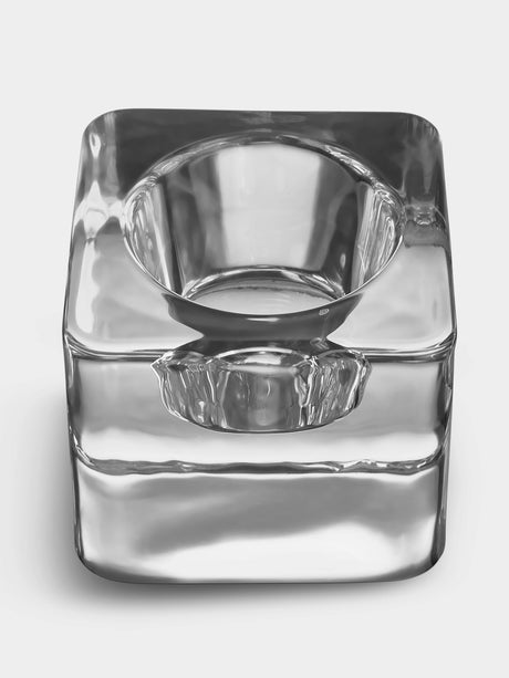 The Orrefors Ice Cube Votive Candle Holder by Goran Warff features a clear, square crystal design with rounded edges and a central circular depression, reminiscent of an ashtray, shown against a plain white background.