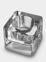 The Orrefors Ice Cube Votive Candle Holder by Goran Warff features a clear crystal square base with a circular center, designed to elegantly hold tealight candles or serve as a stylish ashtray with grooves for resting cigarettes.