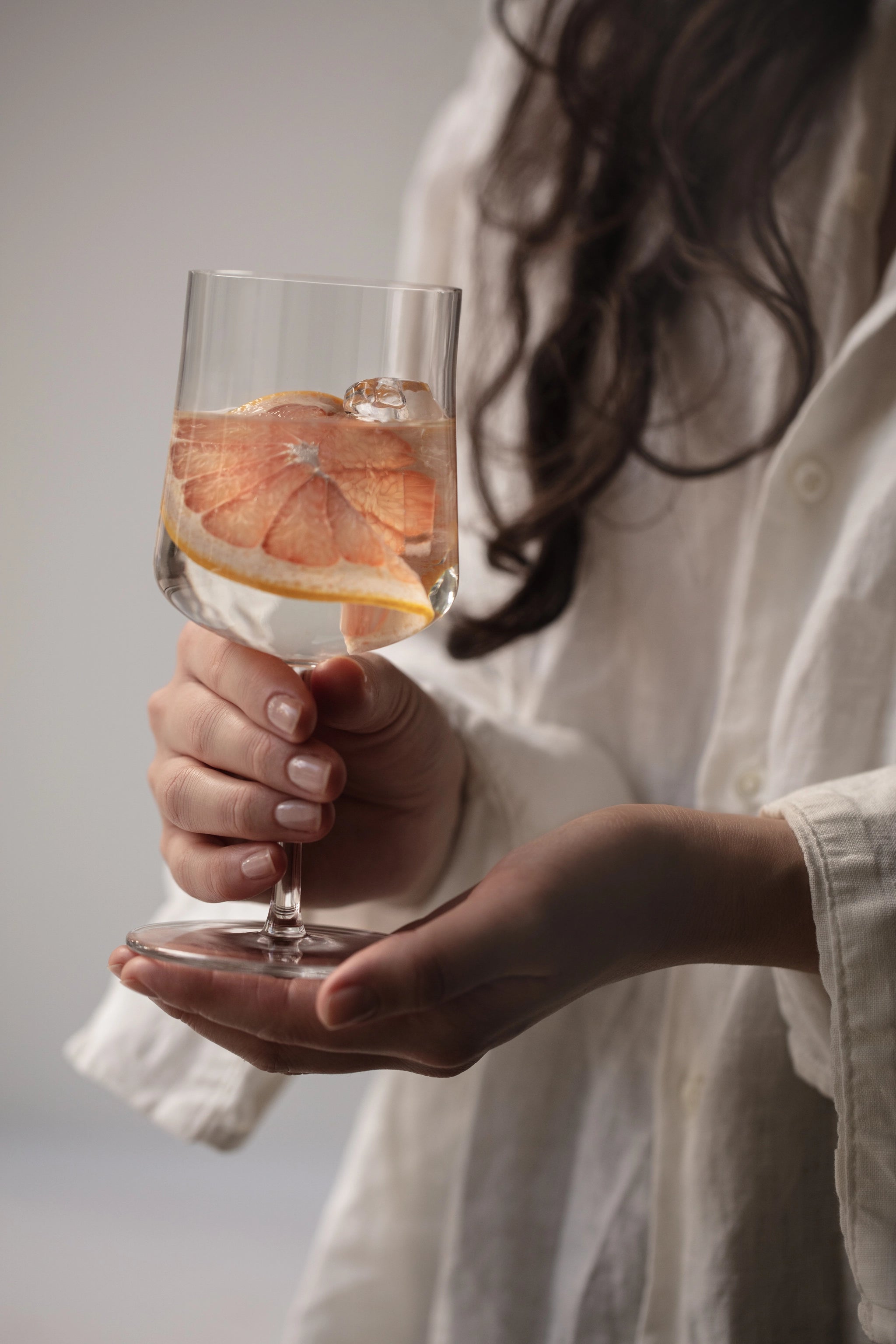 A person in a white shirt holds an Orrefors: Informal Medium Glass 12oz filled with a refreshing drink, ice cubes, and grapefruit slices, highlighting the elegance of Scandinavian design.