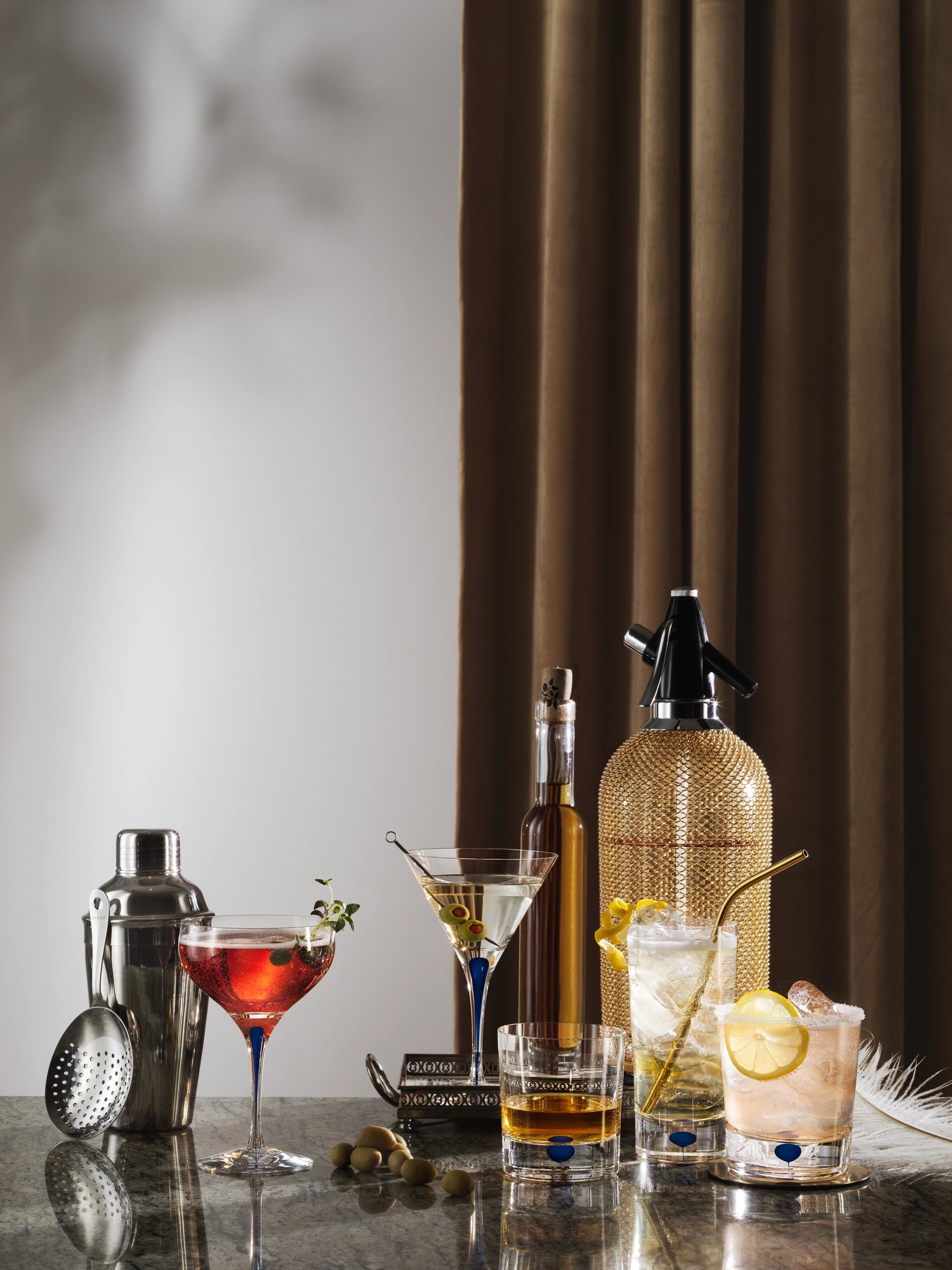 A cocktail setup boasts the elegant 10oz Orrefors Intermezzo Blue Coupe Glass, paired with assorted glasses, a shaker, olives, and a metallic bottle—all perfectly arranged on a reflective surface against a curtain backdrop.