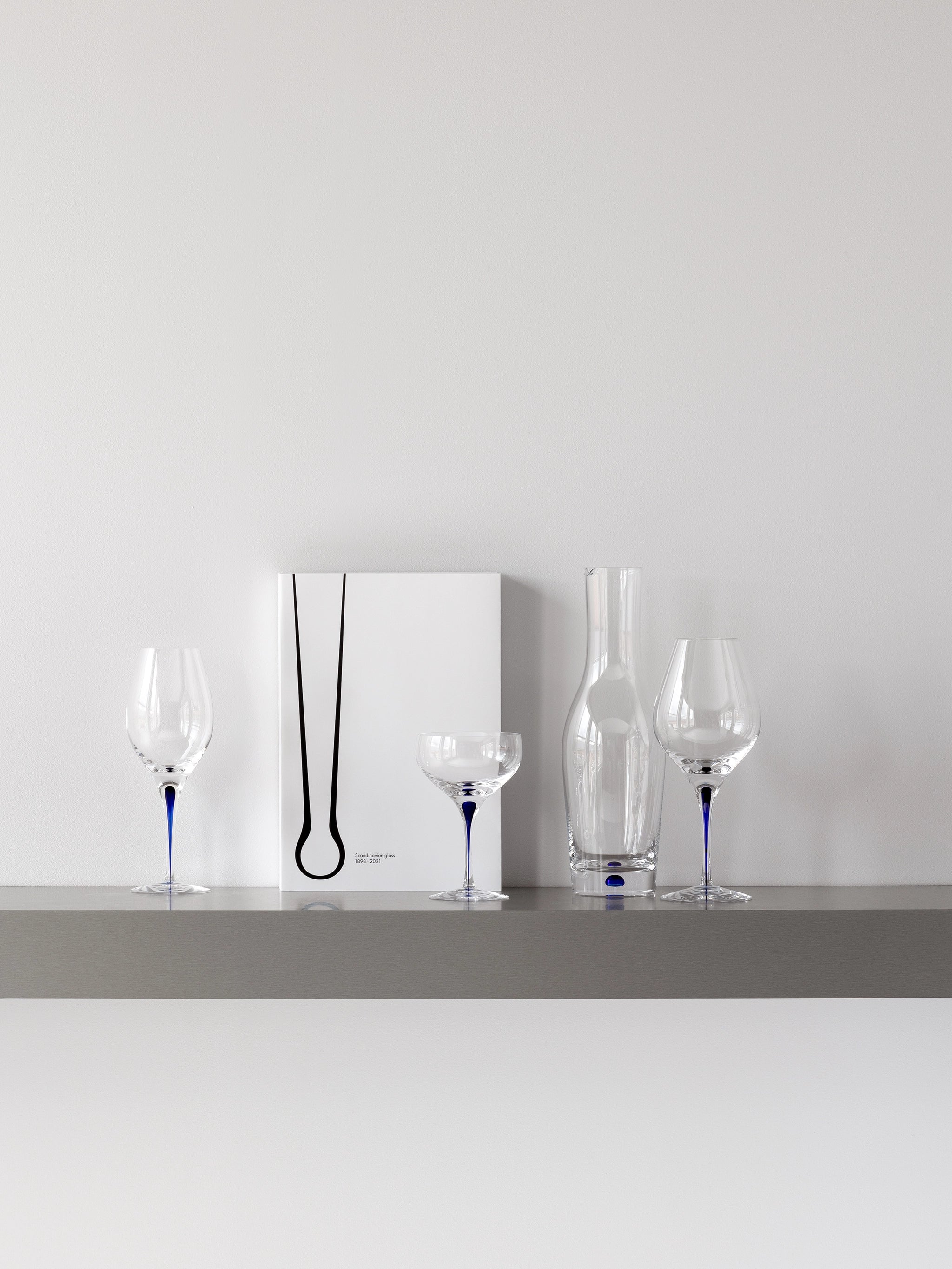 A minimalist shelf displays a white book with a black abstract design, two wine glasses, an Orrefors Intermezzo Blue Coupe Glass 10oz, and a clear glass vase—all with blue accents. Designed by Erika Lagerbielke, these pieces enhance the elegance of any space.