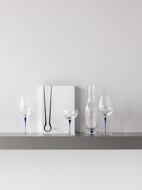 A minimalist shelf displays a white book with a black abstract design, two wine glasses, an Orrefors Intermezzo Blue Coupe Glass 10oz, and a clear glass vase—all with blue accents. Designed by Erika Lagerbielke, these pieces enhance the elegance of any space.