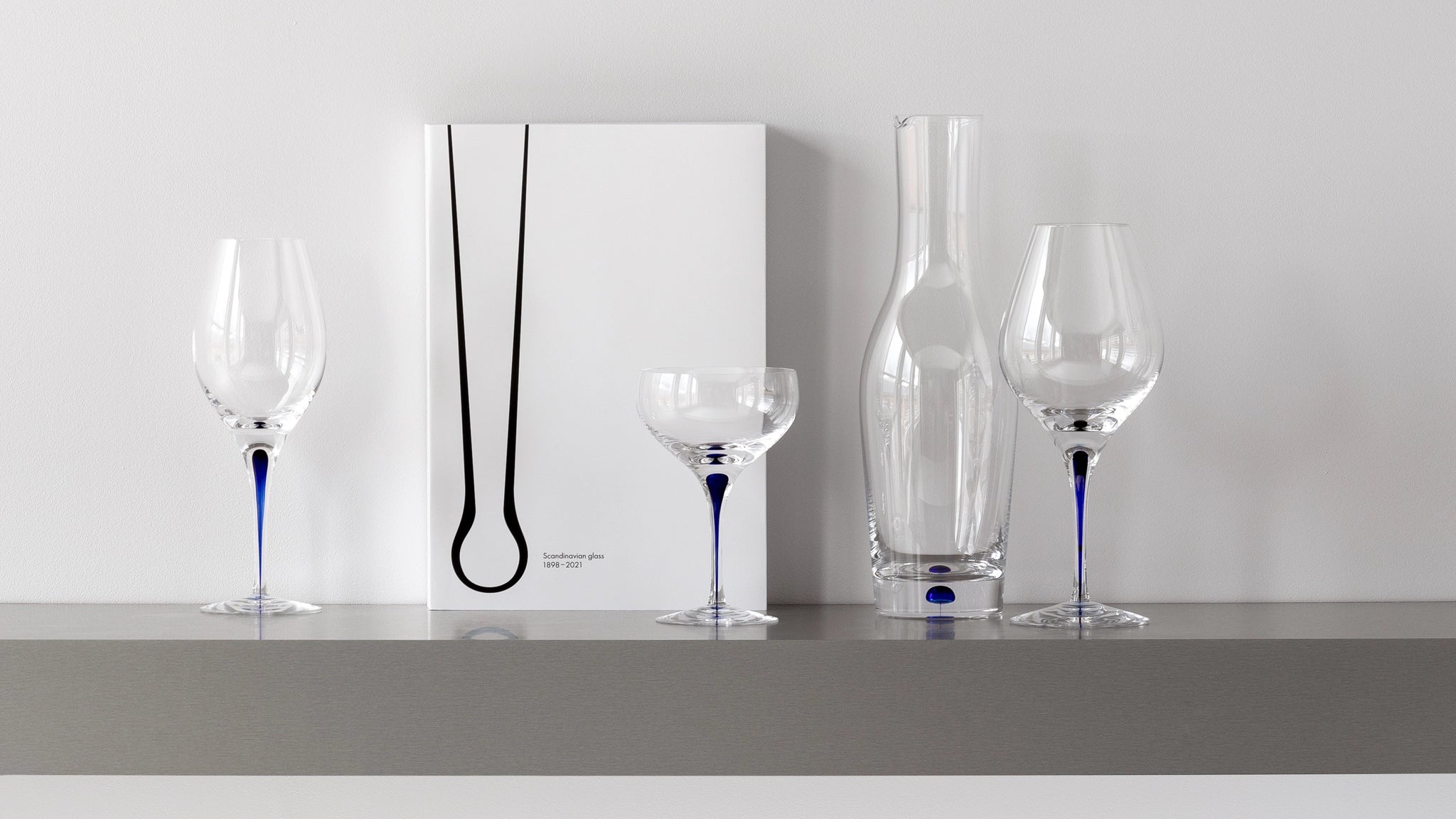 On a shelf sits an elegant collection of Orrefors glassware, including two Intermezzo Blue Bouquet 20oz wine glasses, a cocktail glass by Erika Lagerbielke, a carafe, and a minimalist-design book.