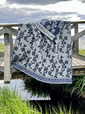 The Blanket: Art Nouveau - Blue - Ullfilt Jugend by Kerstin Landstrom, made from 100% Norwegian lambs wool, drapes over a wooden bridge railing. Below, lush grass and shimmering water under a cloudy sky complete the picturesque scene.