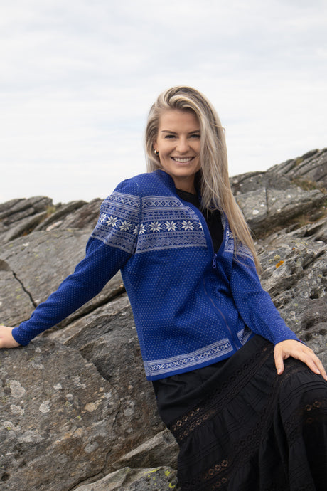 A woman wears the Northern Story Setesdal Royal Blue Norwegian Design Wool Cardigan and a black skirt while sitting on rocky terrain, smiling at the camera under a cloudy sky.