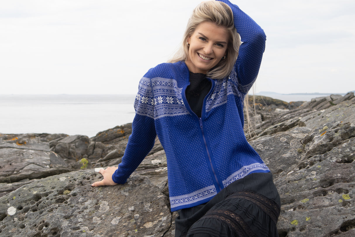 A woman in the Northern Story Setesdal Royal Blue Norwegian Design Wool Cardigan sits on rocky seaside terrain, smiling and resting her left hand on her head.