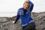 A woman in the Northern Story Setesdal Royal Blue Norwegian Design Wool Cardigan sits on rocky seaside terrain, smiling and resting her left hand on her head.