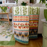The Kustland - Coastal Land 100% Norwegian Lamb's Wool Blanket by Kerstin Landstrom Sweden—OEKO-TEX certified with green, orange, red, blue, and black patterns—lies on a light-colored sofa in a cozy living room.
