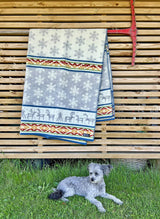 A dog rests on grass before a wooden slatted wall adorned with a snowflake and reindeer-covered Laponia blanket by Kerstin Landstrom. Crafted from OEKO-TEX-certified 100% Norwegian Lambs Wool, it complements the red tool mounted above.