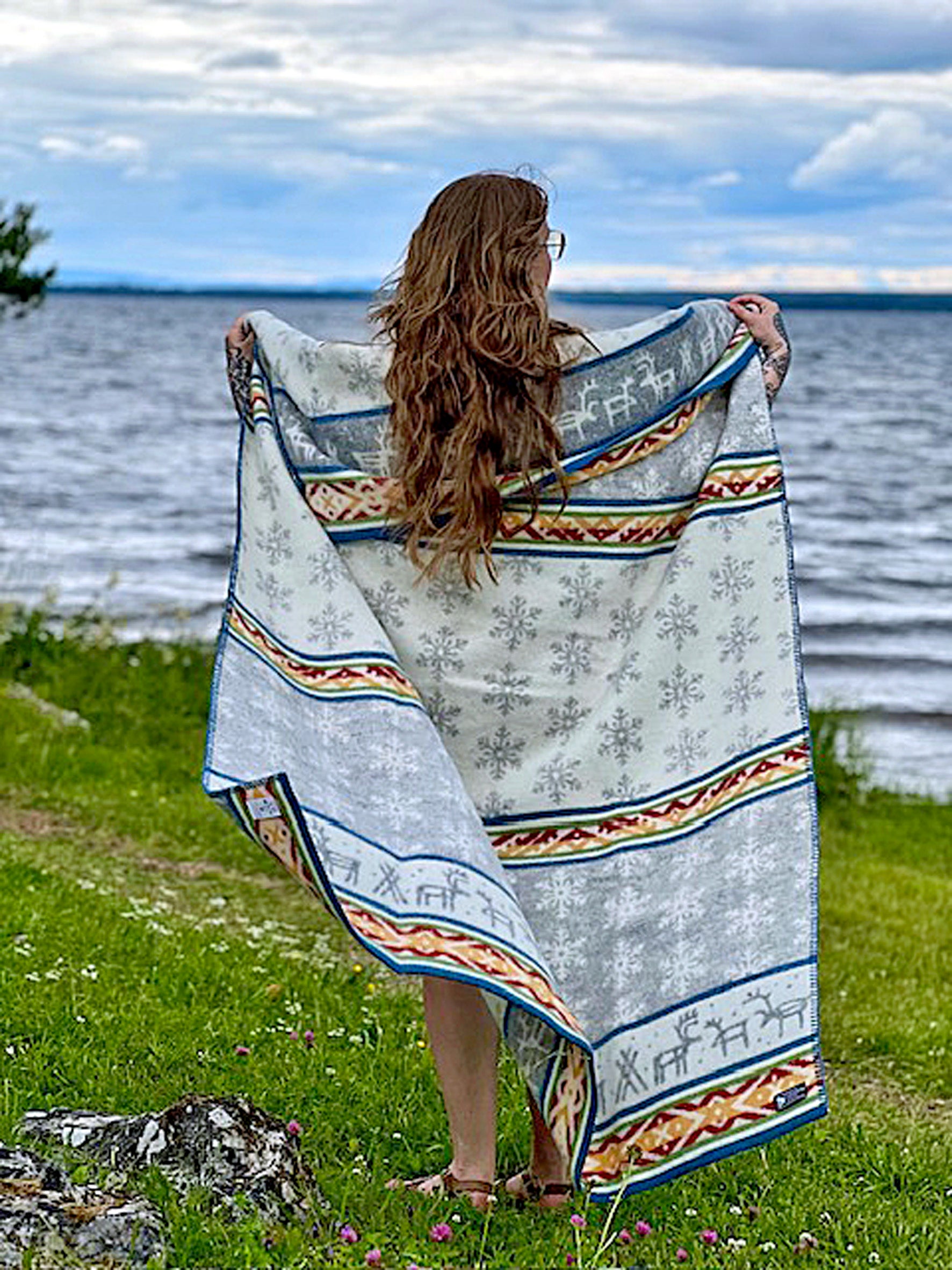 A woman with long hair stands by a lake, holding her outstretched Sámi-inspired blanket Laponia by Kerstin Landstrom, crafted from 100% Norwegian lambs wool and OEKO-TEX certified.