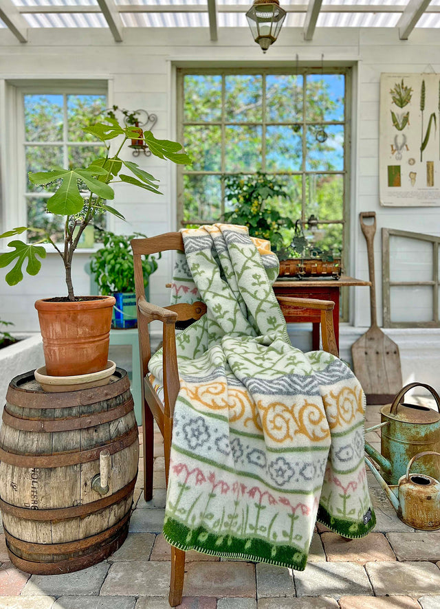 A cozy room with a chair draped in the Linnaeus - 100% Norwegian Lambs Wool Blanket by Kerstin Landstrom, surrounded by plants evocative of the Linnaeus Garden, gardening tools, and a wooden barrel, all illuminated by natural sunlight.