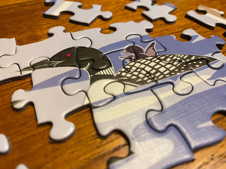 Close-up of a partially completed jigsaw puzzle showing a black and white bird with a red eye, resembling a loon, on a wooden surface.