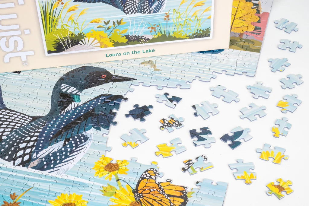 Jigsaw puzzle showing Minnesota loons by a lakeside, with loose pieces on a white surface surrounded by flowers and a butterfly.