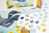 Jigsaw puzzle showing Minnesota loons by a lakeside, with loose pieces on a white surface surrounded by flowers and a butterfly.