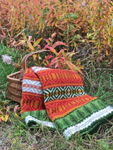 A woven basket rests on grass next to autumn plants, draped with the Mora Blanket—100% Norwegian lamb’s wool by Kerstin Landström Design—in green, orange, white, and red.