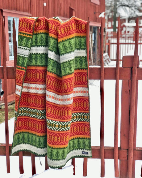 The Mora Blanket—100% Norwegian lamb’s wool by Kerstin Landström Design—featuring red, green, yellow, and white Scandinavian patterns, hangs over a wooden fence with snow and a red building in the background.