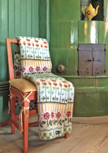 The Blanket: Karin, a 100% Norwegian Lambs Wool creation by Kerstin Landstrom, elegantly drapes over a wooden chair beside a green tiled stove, topped with a teapot, effortlessly combining warmth and style.