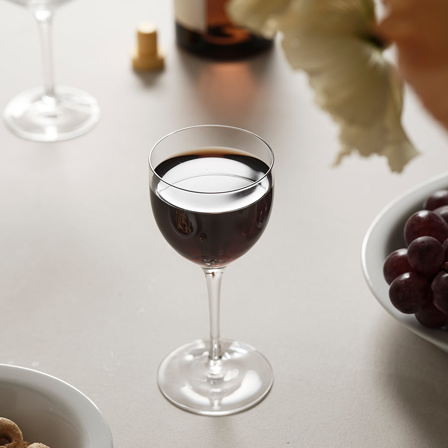 A glass of red wine rests elegantly on the table, surrounded by a bowl of grapes, a wine bottle, and a delicate flower. An Orrefors More Nick & Nora Glass 5.7oz from the 2-Pack adds a touch of sophistication to the scene.