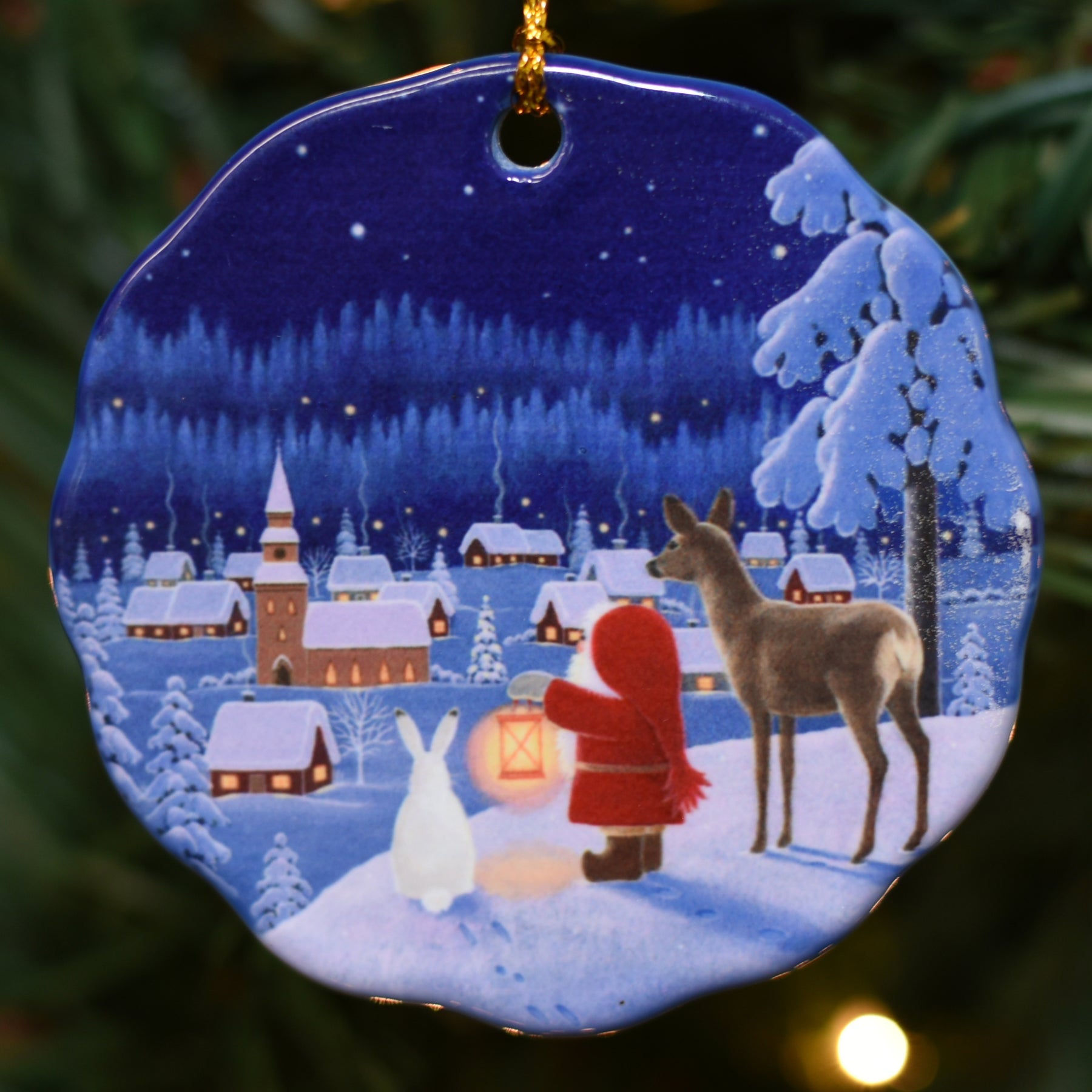 The Ornament: Tomte Reindeer & Rabbit on Porcelain Ornament by Eva Melhuish features a charming winter scene with a child in red, a deer, and a rabbit overlooking a snowy village aglow with lantern lights—a nod to Scandinavian Christmas magic.