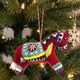 Red felted wool Swedish Dala Horse ornament with colorful embroidery hanging on a Christmas tree, near white snowflake ornament and string lights.