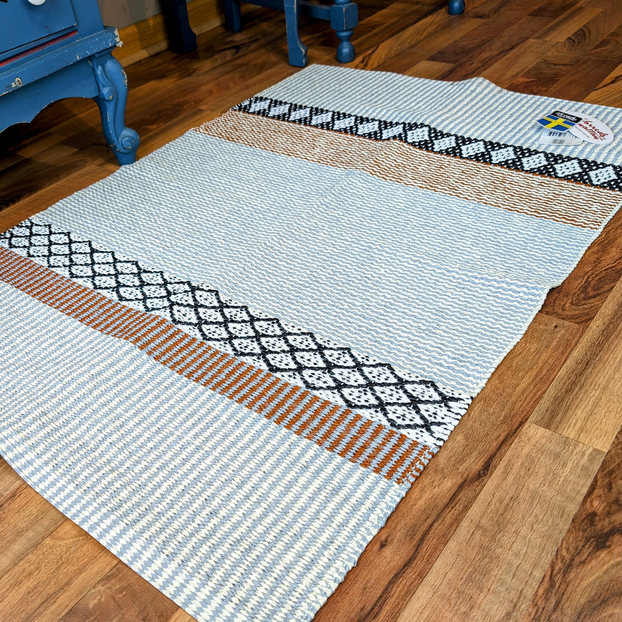 The Savanne Light Grey Vinyl rug, showcasing alternating stripes of blue, white, black, and brown patterns, is elegantly placed on a wooden floor—a testament to Swedish craftsmanship.