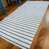 A dark gray woven vinyl rug, Horredsmattan - Music, lies on a wooden floor near blue furniture legs.