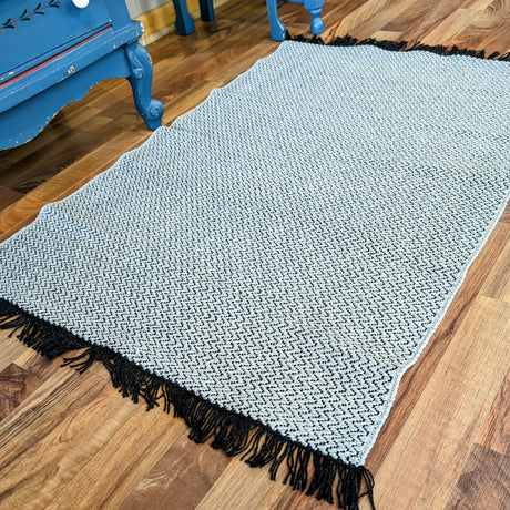 The Horredsmattan - Candy - Gray vinyl rug, rectangular and light gray with a subtle zigzag pattern and black fringed edges, is placed on a wooden floor near blue furniture.