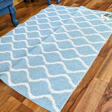 The wooden floor is graced by the Rug: Wave Blue, a Swedish vinyl rug featuring a light blue hue with a white geometric pattern, offering both durability and timeless charm.