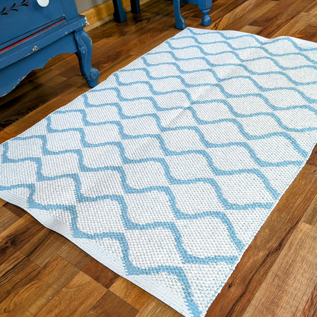 The Wave Blue rug, featuring a durable and long-lasting white weave with a blue lattice pattern, lies on a wooden floor next to a blue piece of furniture.
