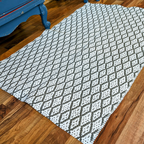 The Eye Grey 70x100cm rug, featuring a durable, maintenance-free woven design with a black and white diamond pattern, complements the wooden floor next to a piece of blue-painted furniture.
