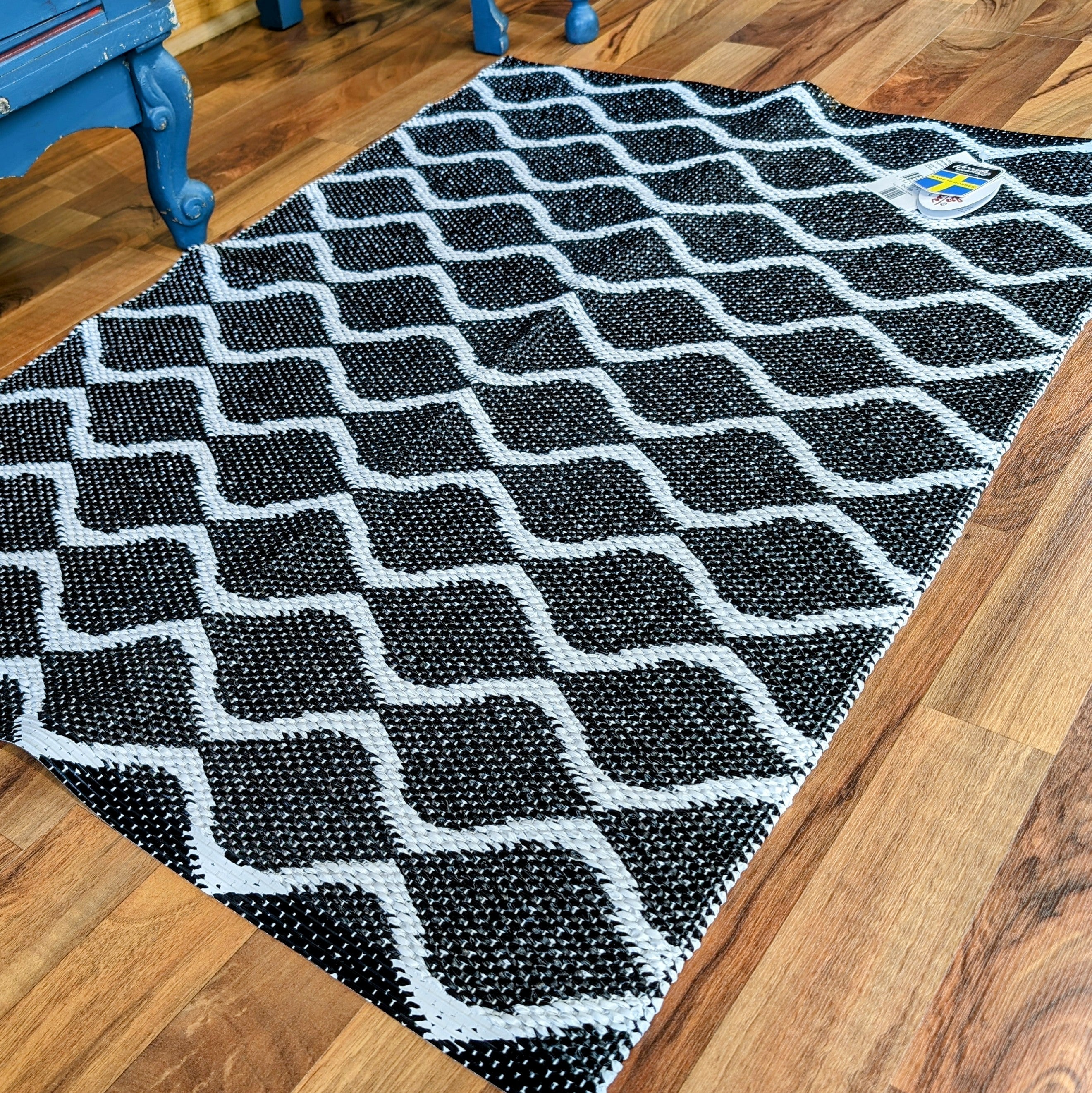 The "Rug: Wave Black" enhances the wooden floor with its reversible black and white geometric pattern, while a blue chair is partially visible in the background. Made from Swedish vinyl, it promises durability and longevity, seamlessly combining style with practicality.