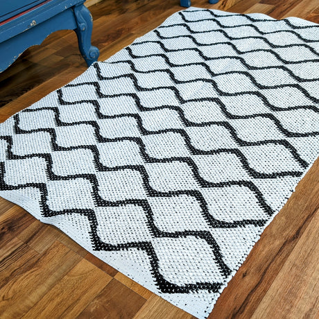 The Wave Black Swedish vinyl rug, featuring a geometric diamond pattern in black and white, is elegantly placed on a wooden floor beside a piece of blue furniture, offering a blend of style and durability that ensures its long-lasting appeal.