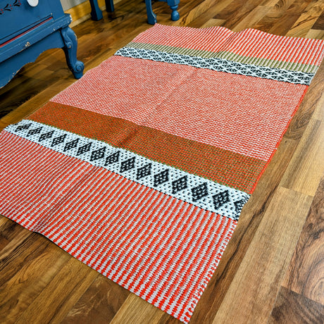 A Horredsmattan Savanne vinyl rug in reddish orange, beige, white, black, and green geometric patterns lies on a wooden floor beside blue furniture.