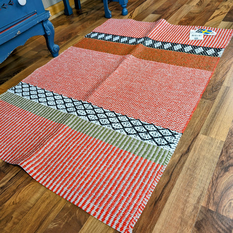 The Horredsmattan Savanne vinyl rug in reddish orange, beige, white, black, and green geometric patterns lies on a wooden floor by a blue piece of furniture. A price tag is attached to one corner of this machine washable rug.