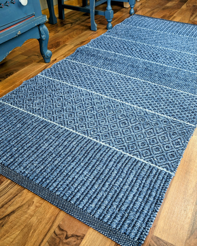 A Mixed Alice rug in blue, made of cotton and vinyl, featuring a textured Swedish design with a geometric pattern, lies on a wooden floor near blue furniture, making it ideal for both indoor and outdoor settings.
