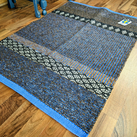 The Cotton/Vinyl Rug: Horredsmattan - Mixed Forest - Cobalt, a machine-washable rug with blue edges and black, white, and blue patterns, lies on a wooden floor. A small patch with a yellow and blue shield appears in one corner.