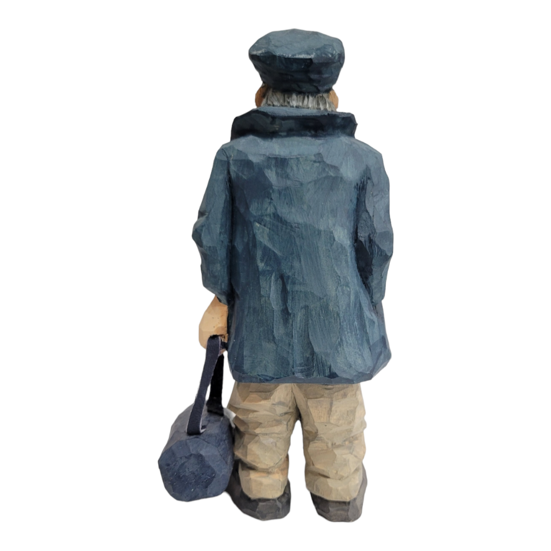 The "Home Port" figurine by Bill Erickson is a hand-carved wooden sculpture depicting a person wearing a cap and blue coat, holding a bag and facing away.