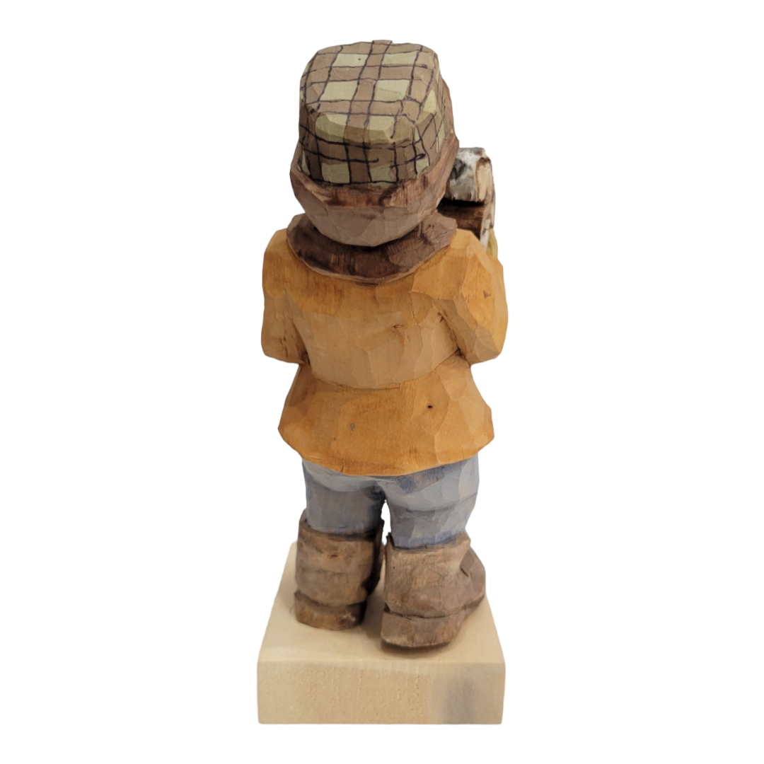 The "Bringing the Firewood" wooden figurine by Bill Erickson showcases a person in a plaid hat and orange coat, standing on a square base, as seen from the back.