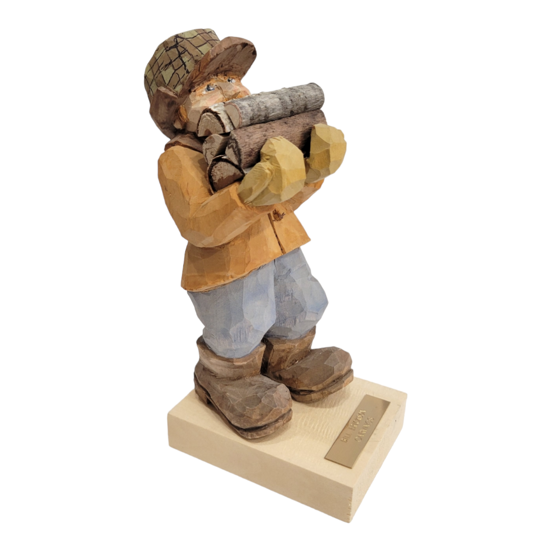 The "Bringing the Firewood" figurine by Bill Erickson is a hand-carved wooden sculpture featuring a person in a cap and boots, holding logs and standing proudly on a rectangular base.