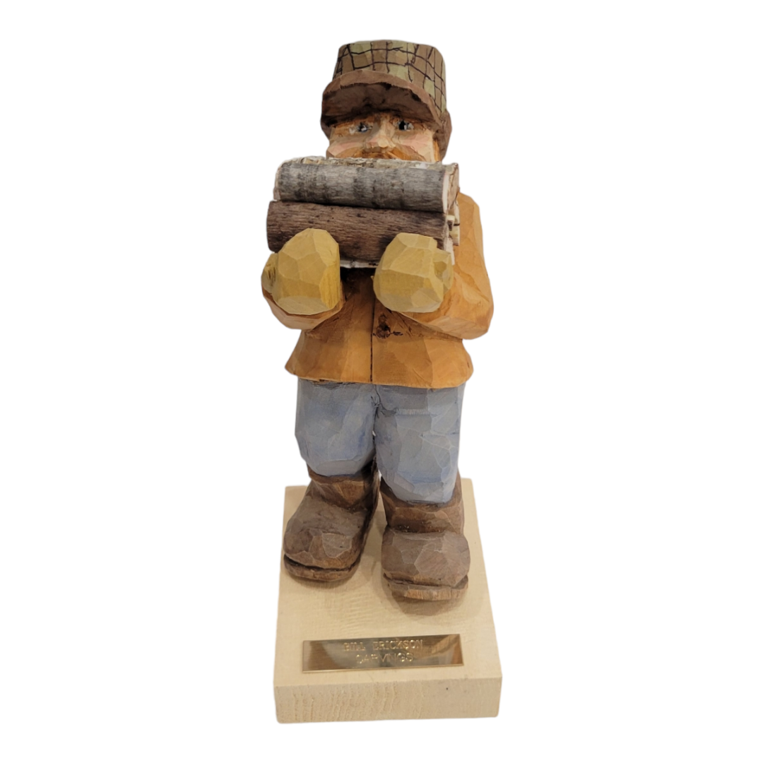 The "Bringing the Firewood" figurine by Bill Erickson is a beautifully hand-carved wooden piece featuring a person in a cap and jacket, holding a log. It stands confidently on a base with an engraved plaque, highlighting exceptional craftsmanship.