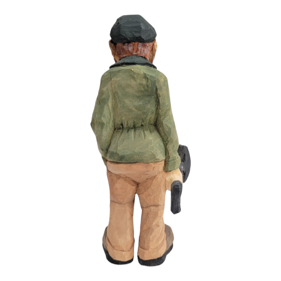 This hand-carved wooden figure of a person, exemplifying Bill Erickson's signature craftsmanship, is dressed in a green coat and cap. From behind, the intricate detail shows them holding a shovel with precision.